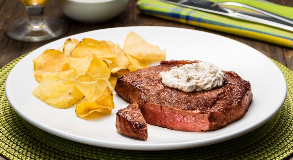 Fatia de contrfilé servido com patê de azeitonas e batatas chips em um prato branco redondo, por cima de um jogo americano verde.