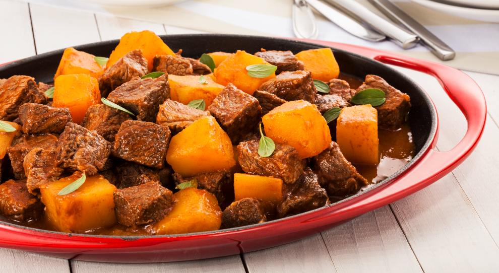 Picadinho de carne bom abóbora servido em um travessa vermelha, em uma mesa de madeira branca.