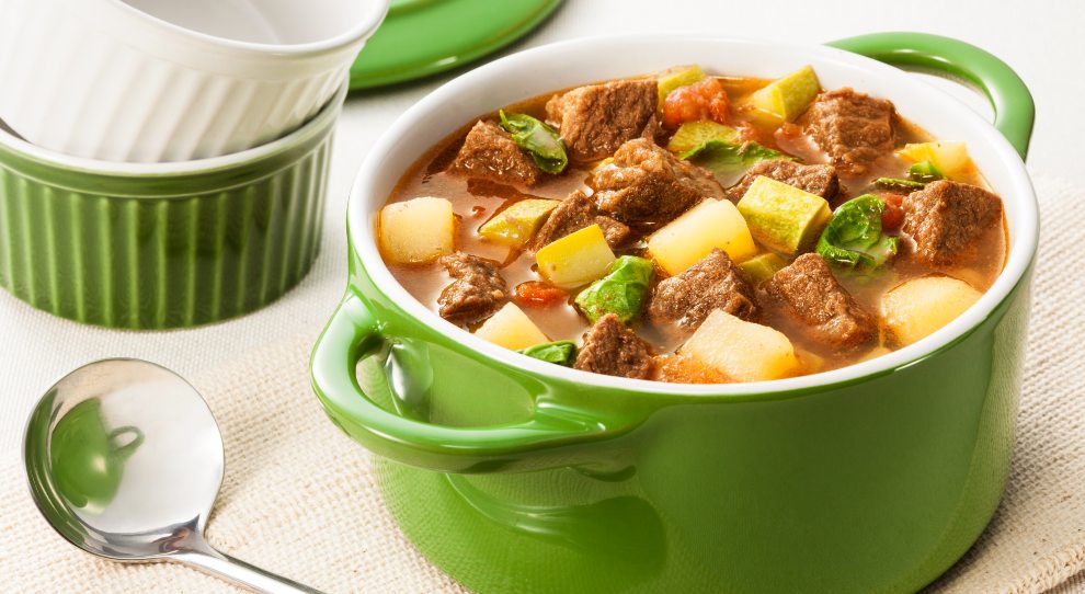 Sopa de carne prepararda com Coxão Duro Friboi e legumes, servida em uma penela de porcelana com o interior branco e de cor verde do lado de fora. Atrás, dois ramquins empilhados, o de baixo na com verde e o de cima na cor branca. Ao lado da panela, à esquerda, um conxa prateada.