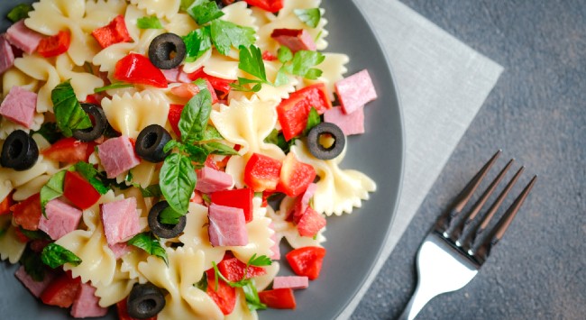 Salada de macarrão com azeitona, manjericão, tomate e cubos de tender em um prato cinza.