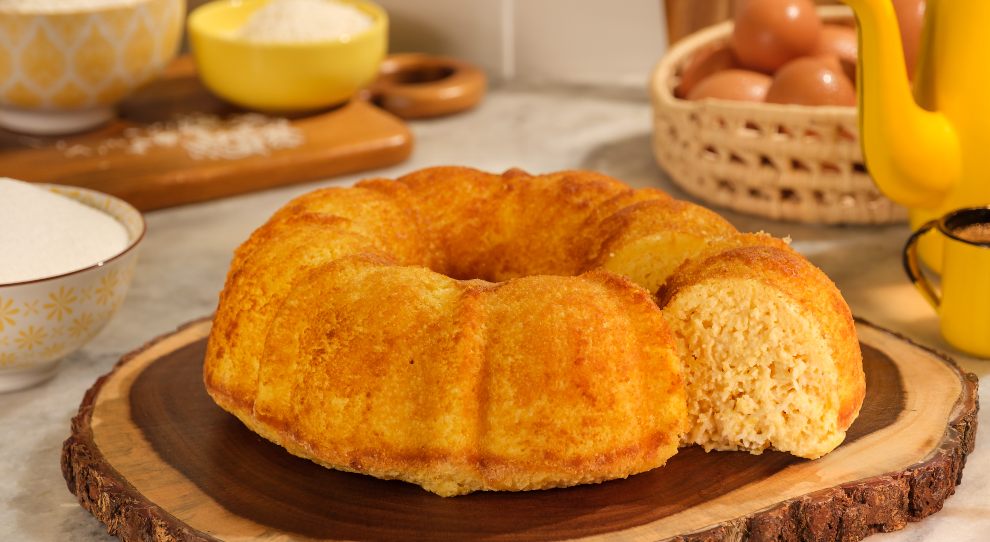 Bolo de milho cremoso circular em uma tábua de madeira rústica parecida com tronco de árvore. Ao lado direito, uma garrafa de café e uma xícara, ambas amarelas e esmaltadas. Atrás da garrafa, uma cesta de sisal com ovos caipiras. À esquerda da cesta, uma tábua de madeira grossa com tigelas de farinha de trigo. À frente, ao lado esquerdo do bolo, uma tigela com açúcar refinado.