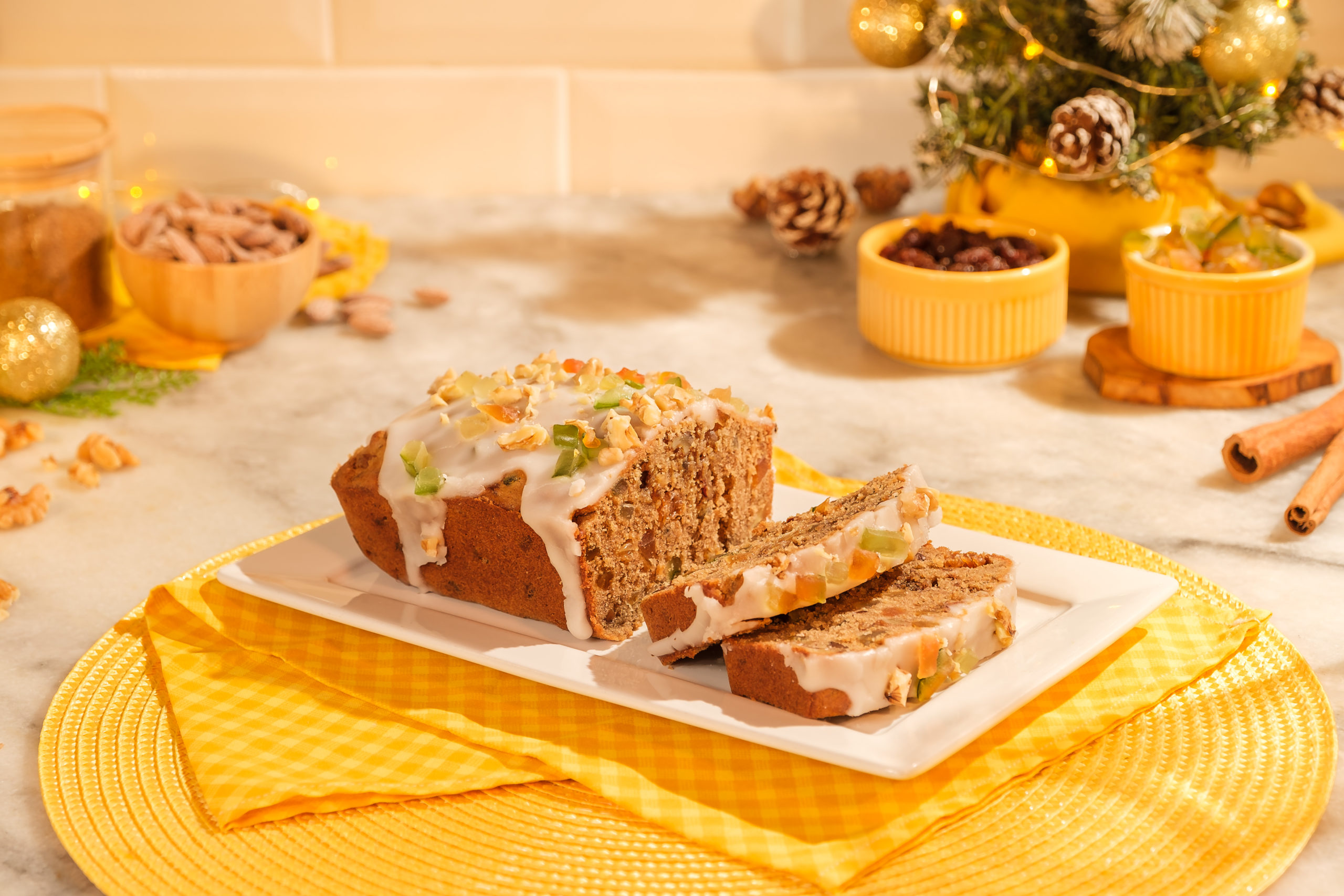 Bolo de natal em formato retangular, servido em fatias em um prato branco retangular. Por cima do bolo há uma cobertura branca e frutas cristalizadas.