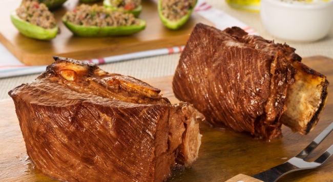 Churrasco de costela servido em cima de uma tábua de madeira. Ao fundo da foto, abobrinha recheadas com carne.