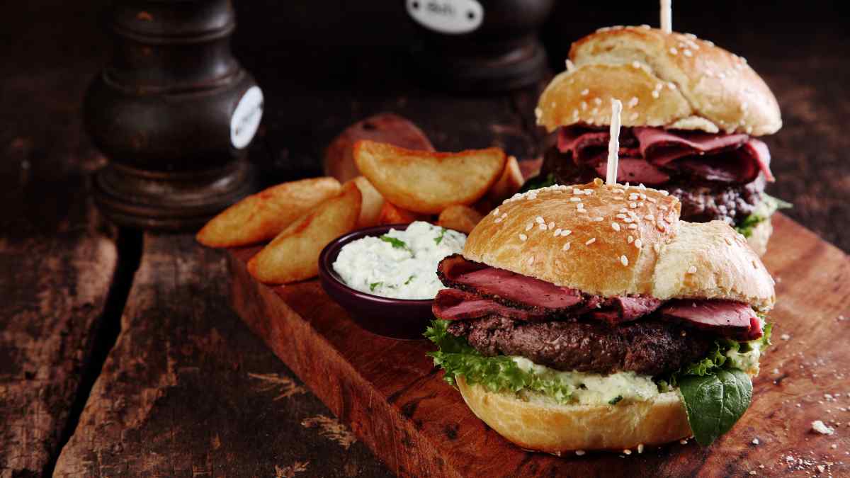 Hambúrguer depastrami servido no pão com gergelim com alface e maionese, em uma tábua de madeira rústica companhado de batata canoa frita.
