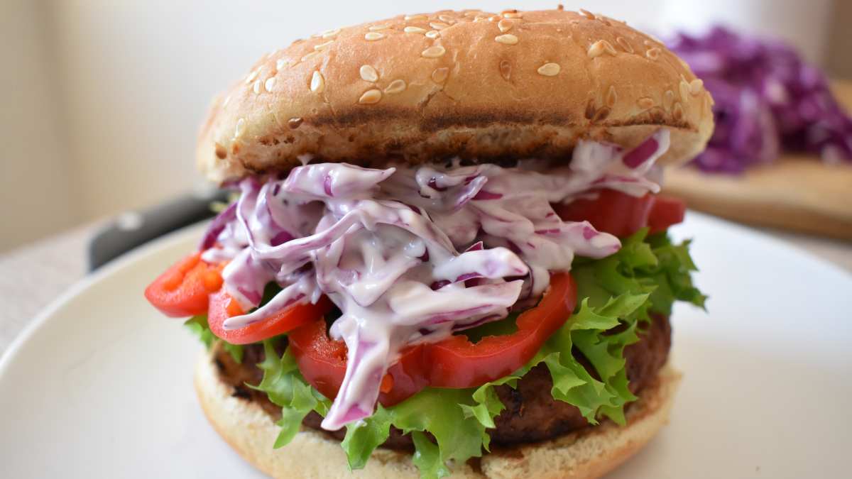 Hambúrguer preparado com salada coleslaw, fodelas de pimentão vermelho e alface no pão com gergelim em um prato branco raso.