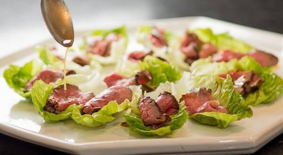 A imagem mostra uma salada de rosbife cuidadosamente montada em um prato branco. No centro do prato, há uma porção generosa de rosbife fatiado em camadas finas, disposto de maneira circular. O rosbife é de cor avermelhada e tem uma aparência suculenta. Ao redor do rosbife, estão dispostos ingredientes coloridos que complementam a salada, como folhas verdes frescas, tomates cereja vermelhos e amarelos, e fatias finas de pepino verde. As folhas verdes estão dispostas de maneira harmoniosa, proporcionando contraste e textura ao prato. A salada é finalizada com um leve tempero, visível nas folhas e no rosbife. O prato está sobre uma superfície branca neutra, destacando os ingredientes coloridos da salada.