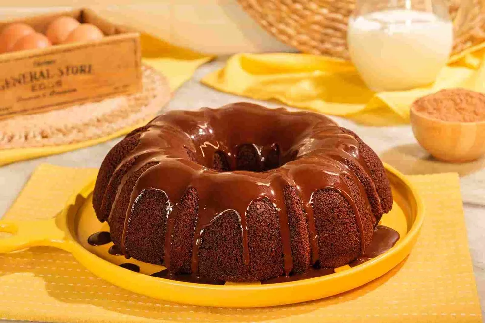 "Um bolo de chocolate úmido e delicioso, coberto com uma generosa camada de ganache. O bolo está em um prato amarelo sobre uma mesa com uma toalha xadrez. Ao fundo, há uma caixa de ovos de madeira, um jarro de leite e um recipiente com pó de cacau.