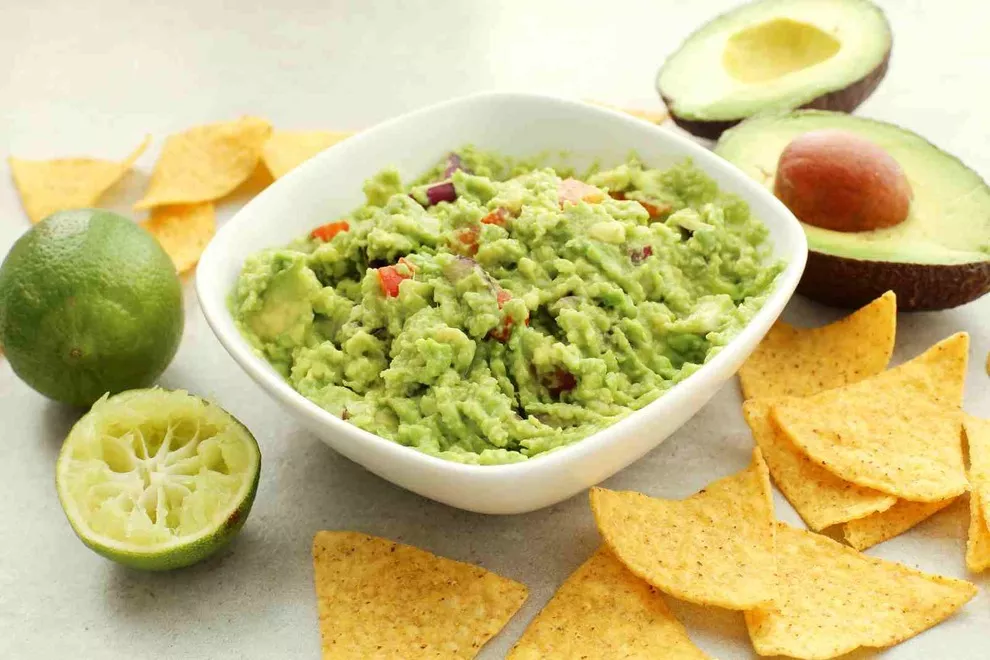 Imagem de uma tigela quadrada branca com Guacamole, sob uma mesa branca com diversos ingredientes ao redor 