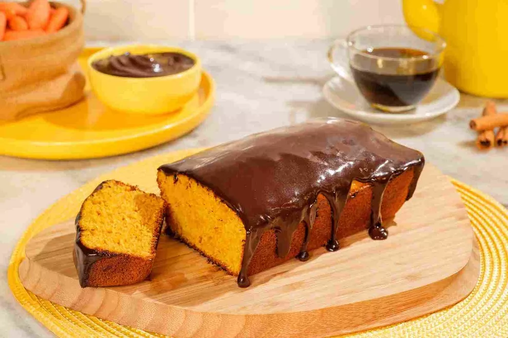 Bolo de cenoura em formato de pão de ló, com glacê de chocolate brilhando. O bolo está apresentado em uma tábua de madeira sobre um jogo americano amarelo. 