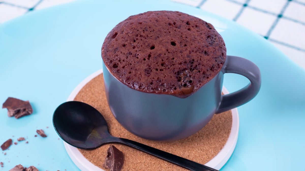 A imagfm destaca um brownie de caneca servido em uma caneca azul, em cima de um suporte redondo com bordas brancas. Do lado, um colher preta.