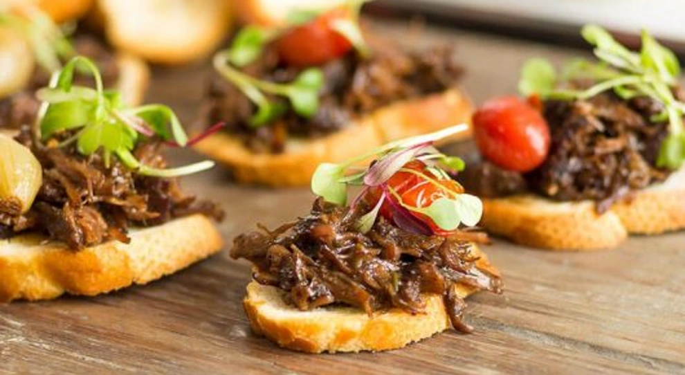 Na imagem, há várias bruschettas sobre uma superfície de madeira. Cada bruschetta é feita com uma fatia de pão baguete coberta com carne desfiada bem temperada. Em cima da carne, há pequenos brotos verdes e fatias de tomate-cereja. O prato apresenta uma aparência fresca e apetitosa, com uma mistura de cores e texturas que destaca os ingredientes utilizados.