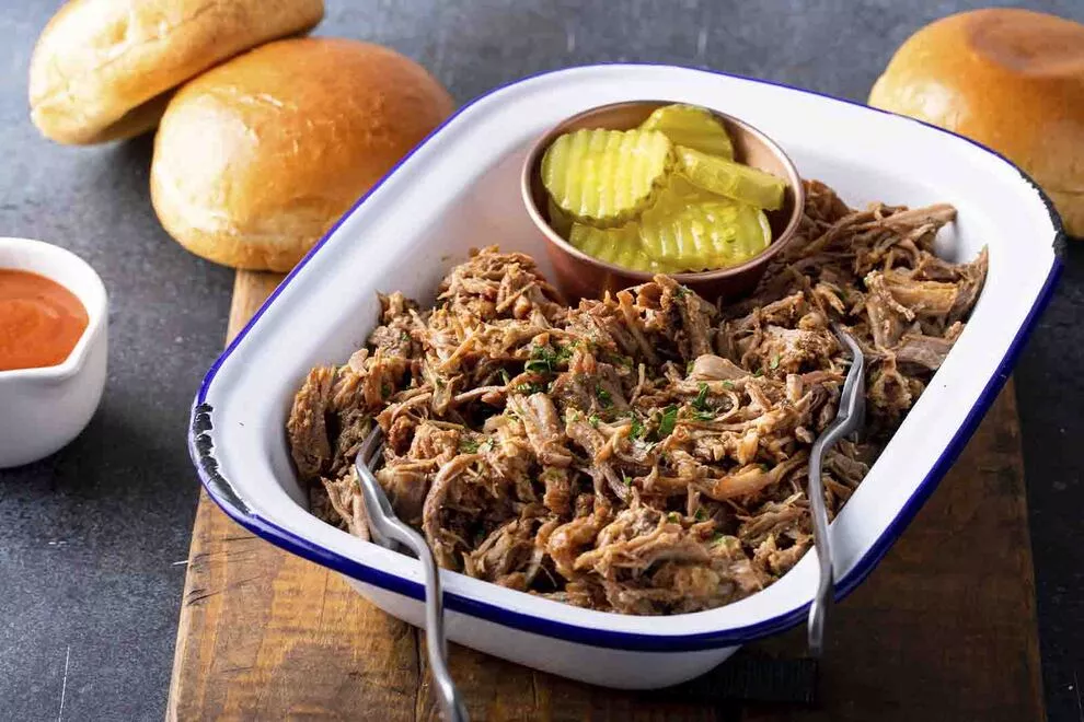 imagem de uma travessa branca retangular com carne louca, com dois garfo em cima dele e alguns ingredientes ao redor, sob uma mesa escura 