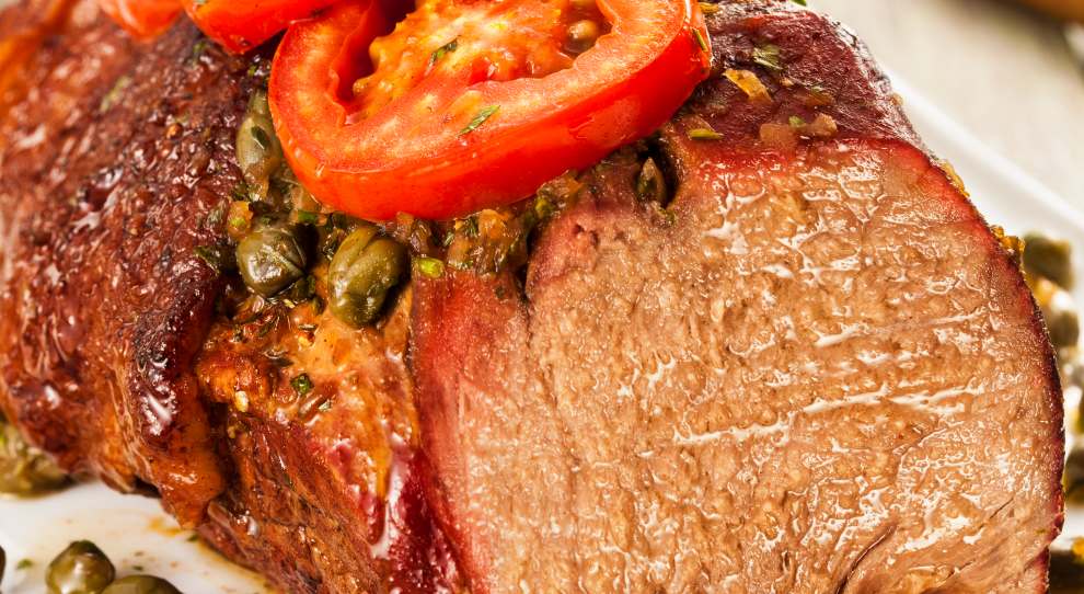 A imagem mostra um prato de coração de alcatra assada. A carne está suculenta e bem cozida, com uma crosta dourada por fora. No topo da carne, há fatias de tomate vermelho e ramos de alecrim, que adicionam cor e frescor ao prato. Ao redor da carne, estão espalhadas alcaparras, que dão um toque de sabor salgado e acidez. O prato está disposto em um prato branco, e o fundo é de uma superfície de madeira, proporcionando um contraste rústico e atraente à apresentação.