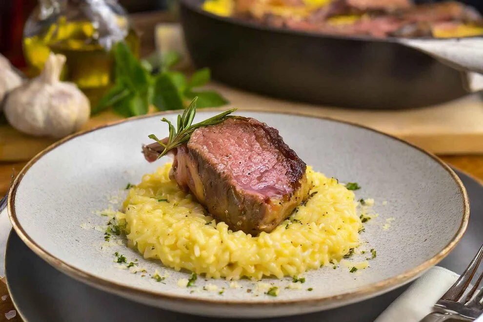 Imagem de uma fatia de cordeiro assado sob um risoto amarelo, em cima de um prato cinza, sob uma mesa de madeira  