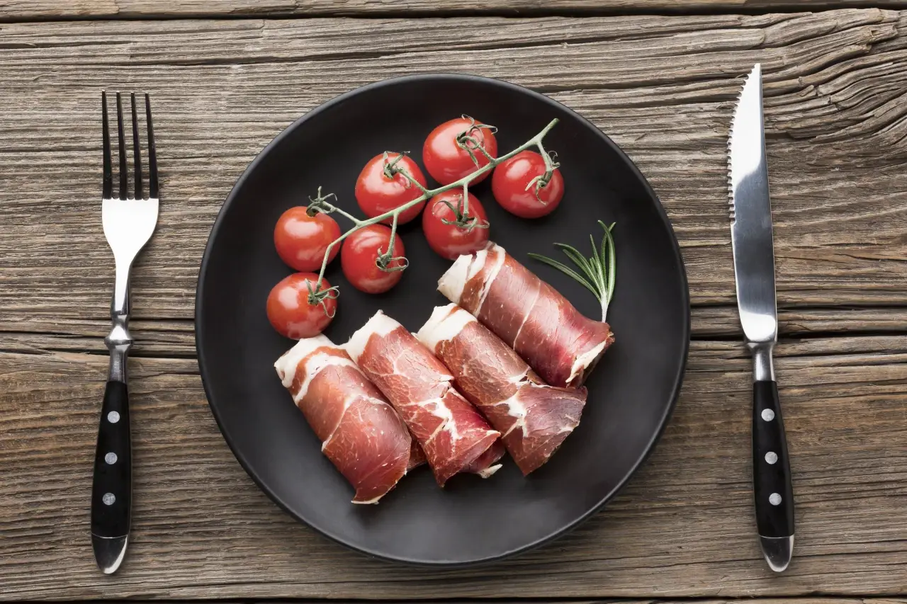 mesa de madeira com prato preto com tomate e rolinho de parma com garfo e faca ao lado do prato