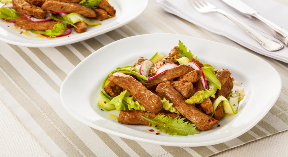 A imagem mostra dois pratos de salada com tiras de carne. A salada é servida em pratos brancos e contém pedaços de carne grelhada, alface, rodelas de rabanete, fatias finas de pepino e folhas de aipo. As tiras de carne são marrons e suculentas, dispostas sobre uma cama de vegetais frescos e crocantes. No fundo da imagem, há um segundo prato de salada semelhante, e ao lado dele estão um garfo e uma faca de prata sobre guardanapos brancos dobrados. A mesa tem um forro com listras horizontais em tons de bege e branco, criando um visual elegante e apetitoso.