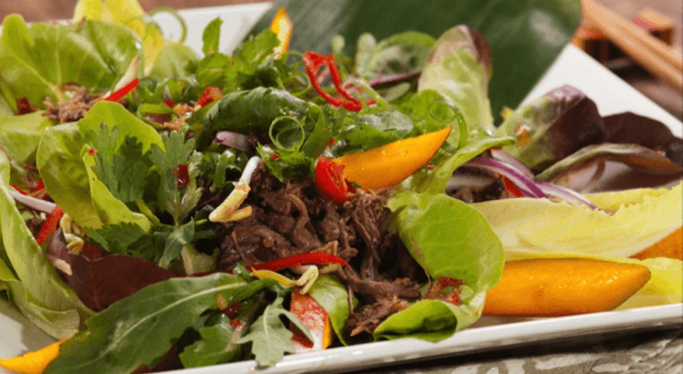 A imagem mostra um prato de salada tailandesa que inclui músculo bovino desfiado. A salada é vibrante e colorida, com uma mistura de folhas verdes frescas e variadas, incluindo alface e outras verduras. Entre as folhas, há pedaços de manga amarelos, que adicionam um toque de cor e provavelmente um sabor doce à salada.

Outros ingredientes visíveis incluem fatias finas de cebola roxa, tiras de pimentão vermelho, coentro fresco e alguns brotos. O prato é apresentado de forma atraente em um prato branco, com os ingredientes distribuídos uniformemente, criando um visual apetitoso e harmonioso.

A combinação de ingredientes sugere um prato equilibrado, com uma mistura de sabores frescos, doces, e picantes típicos da culinária tailandesa.