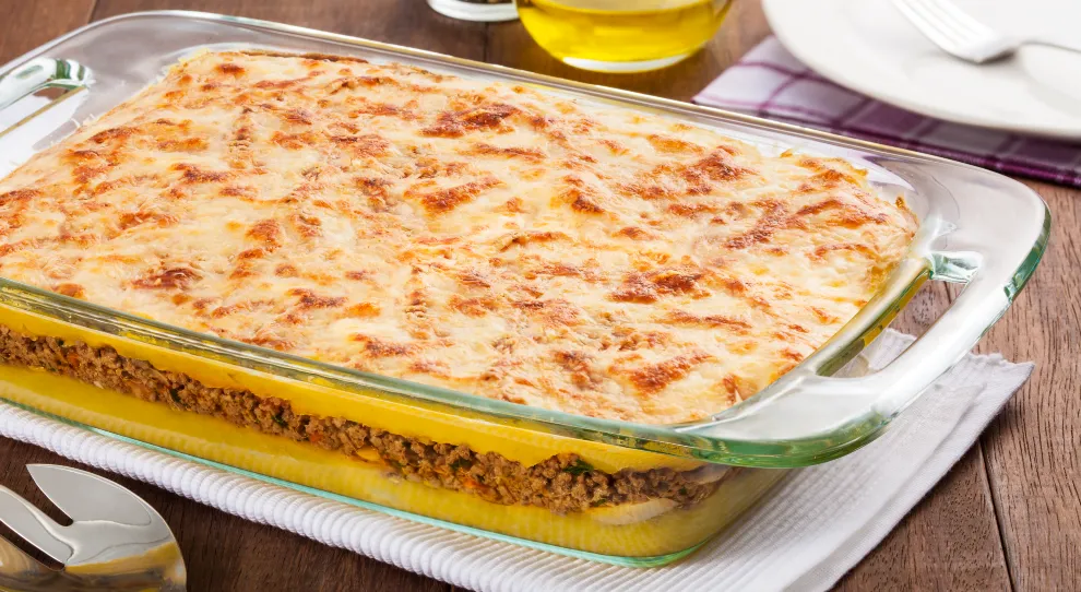 Na imagem, há um prato de vidro retangular contendo uma torta madalena recém-saída do forno. A torta tem uma camada superior dourada e gratinada de queijo, com manchas marrom-douradas que indicam que o queijo foi derretido e gratinado até ficar crocante.

Abaixo da camada de queijo, é possível ver uma camada de massa amarela e, logo abaixo, uma camada de carne moída bem temperada, misturada com pequenos pedaços de vegetais. O prato está sobre um pano de prato branco listrado, colocado sobre uma mesa de madeira rústica.

No fundo da imagem, parcialmente desfocados, há um recipiente com azeite de oliva, um pequeno pote de vidro com pimenta-do-reino em grãos, e um conjunto de pratos brancos com talheres prateados sobre um guardanapo roxo. Uma colher de servir prateada está posicionada à frente do prato.