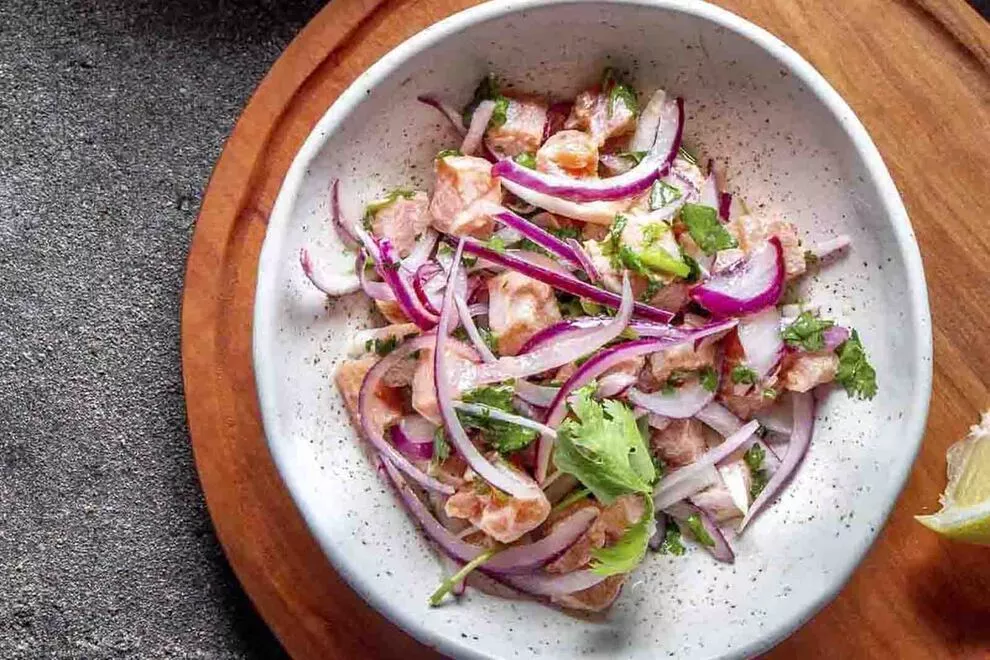 Imagem de um Ceviche de salmão em cima de um prato redondo branco de cerâmica, em cima de uma tábua de madeira redonda, sob uma mesa cinza, visto de cima   