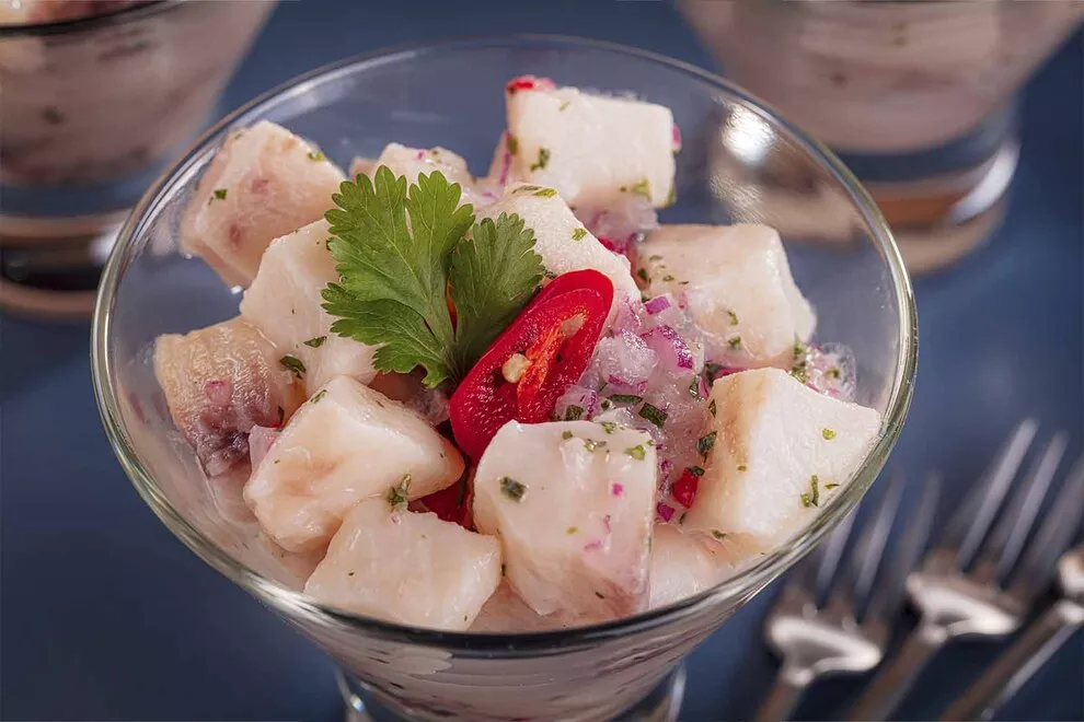 Imagem de um Ceviche de tilápia, em cima de uma taça de vidro, com alguns garfos ao lado, sob uma mesa azul 