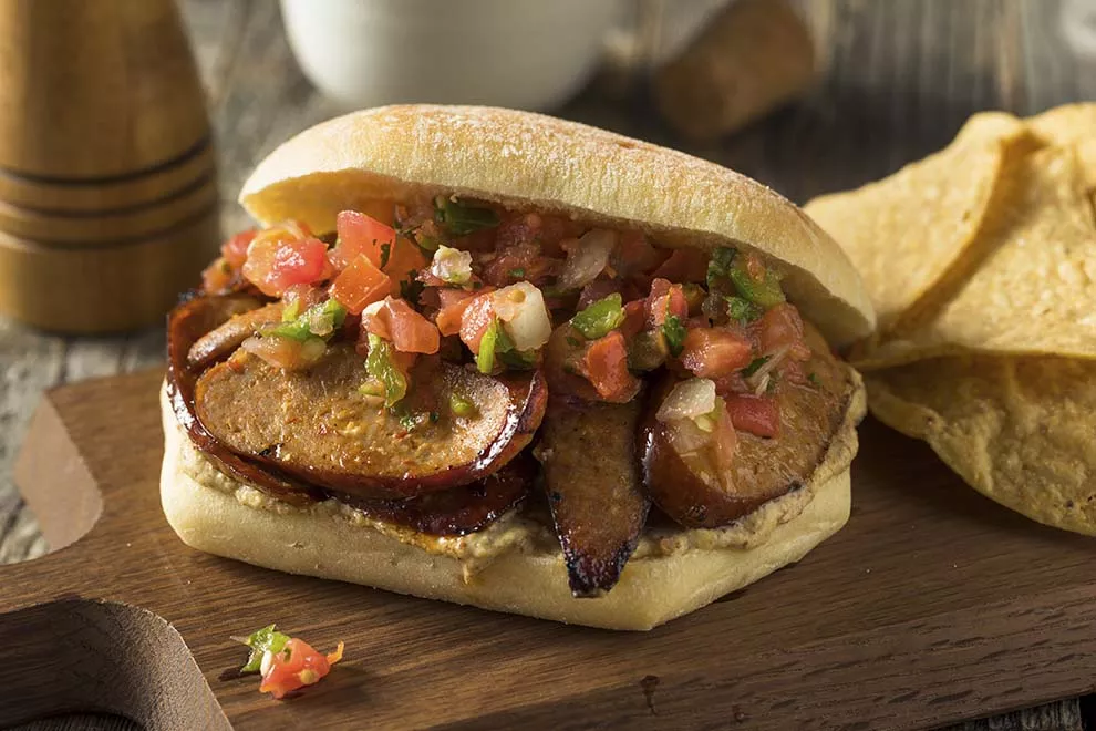 Imagem de uma unidade de choripan, em cima de uma tábua de madeira, com algumas unidades de nachos ao lado, sob uma mesa de madeira 