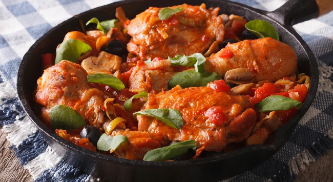 A imagem mostra uma frigideira de ferro cheia de filés de sobrecoxa cozidos em um molho vermelho, preparado à base de tomate. Os pedaços de frango estão cobertos com ervas frescas, como manjericão, e rodeados por legumes como alho e cebola. A frigideira está posicionada sobre um pano com estampa xadrez em tons de azul e branco, que está sobre uma mesa rústica de madeira. O prato tem uma aparência suculenta e bem temperada, transmitindo uma sensação de comida caseira e reconfortante.