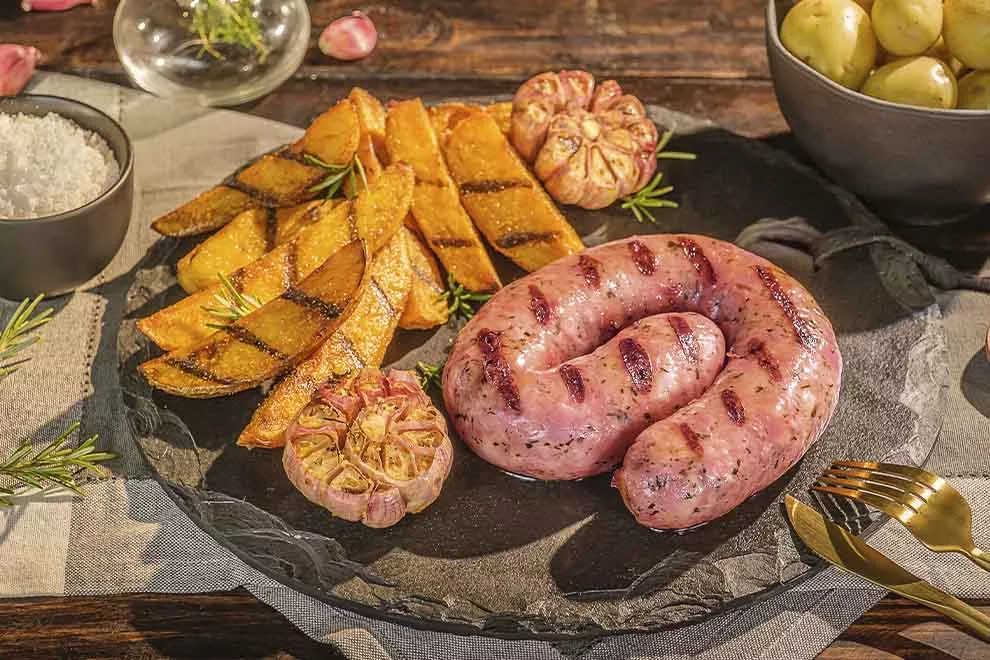 Imagem de uma unidade de linguiça cuibana, com batatas assadas e cabeças de alho assados ao lado, sob um prato redondo de granito, com um par de talheres ao lado, com diversos ingredientes ao redor, em cima deum pano cinza dobrado, em cima de uma mesa de madeira escura 