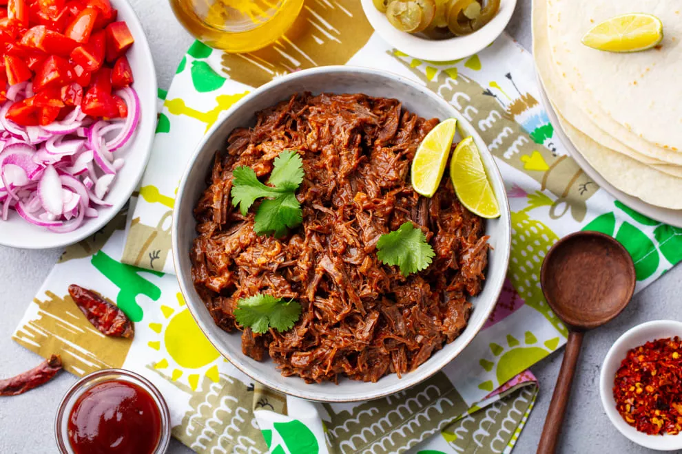 Carne desfiada mexicana servida em uma tigela com coentro e fatias de limão, cercada por ingredientes como tomates picados, cebolas roxas fatiadas, jalapeños, tortillas e flocos de pimenta em uma toalha de mesa colorida, pronta para ser usada em uma refeição tradicional mexicana.