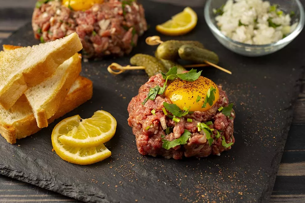 Imagem de um steak tartare com gema de ovo por cima, uma fatia de pão ao lado, com cebolas e salsinha picada por cima, sob uma travessa escura, em cima de uma mesa de mármore 