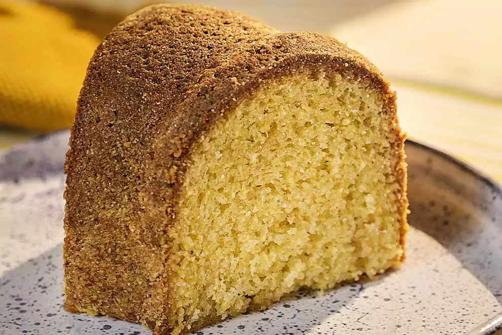 Imagem de uma fatia de bolo de fuba em cima de um prato branco, sob uma mesa de mármore 