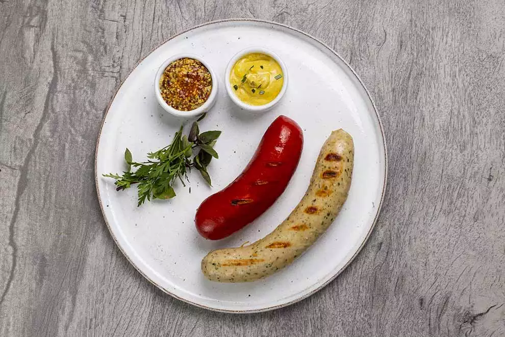 Cervelá com Weisswurst ao lado, sob um prato redondo branco, com duas combucas de molho, em cima de uma mesa de madeira.