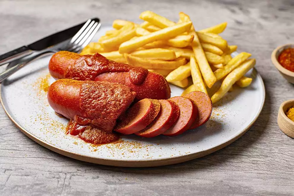 Duas unidades de Currywurst com algumas fatias dele cortadas, com batatas fritas e um par de talheres ao lado, sob um prato redondo, em cima de uma mesa de madeira.