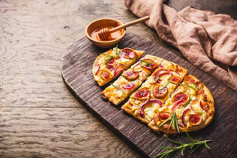 Flammkuchen cortado em em fatias, sob uma tábua de madeira, com uma combuca com mel ao lado, em cima de uma mesa de madeira.