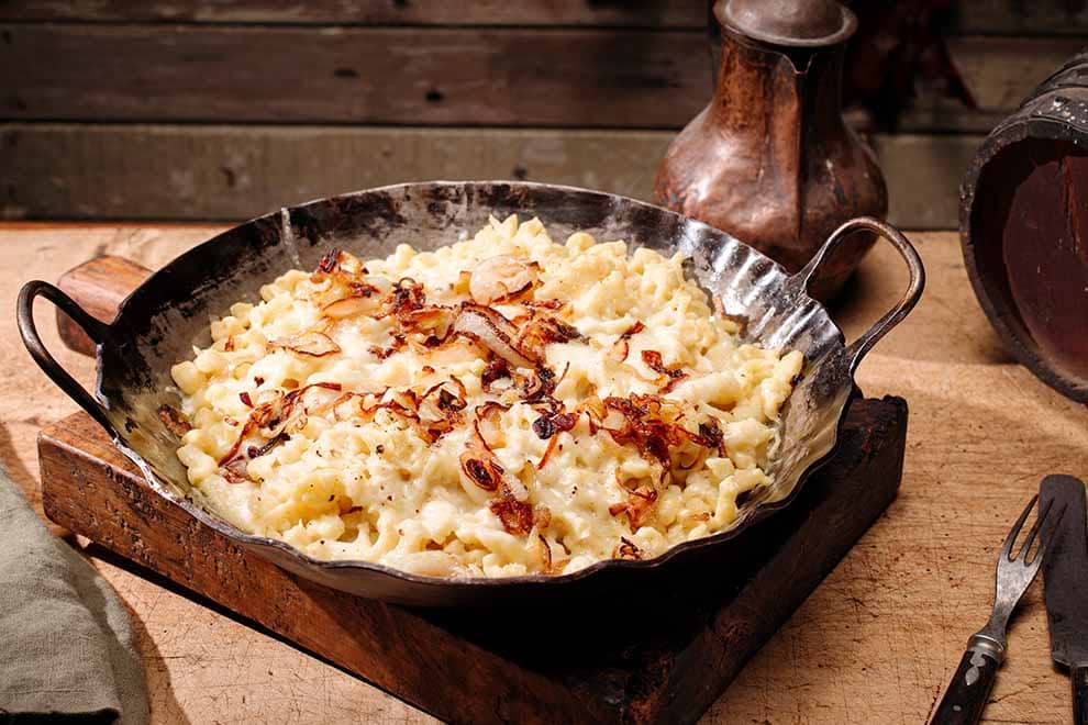 Spätzle em cima de uma panela redonda escura, sob uma tábua de madeira, com um par de talheres ao lado, em cima de uma mesa de madeira.