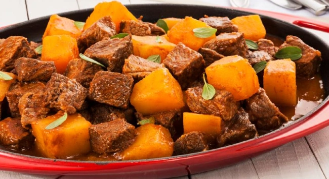 Picadinho de carne bom abóbora servido em um travessa vermelha, em uma mesa de madeira branca.