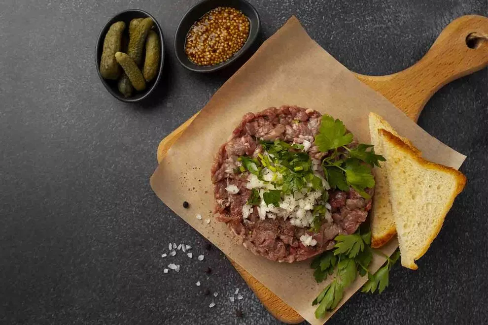 Imagem de uma carne de onça com duas fatias de pão de forma ao lado, com alguns ingredientes ao redor sob uma tábua de cortar carne quadrada, em cima de uma mesa escura 