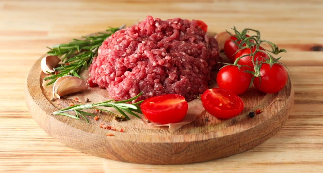 Carne moída em cima de uma tábua de madeira redonda com tomates cereja cortados e ramos de alcrim em volta.