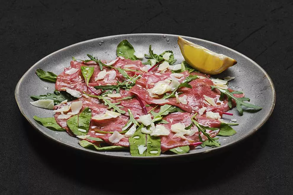 Imagem de um prato escuro com Carpaccio de carne, algumas folhas de rúcula por cima e uma fatia de limão siciliano ao lado, sob uma mesa escura.