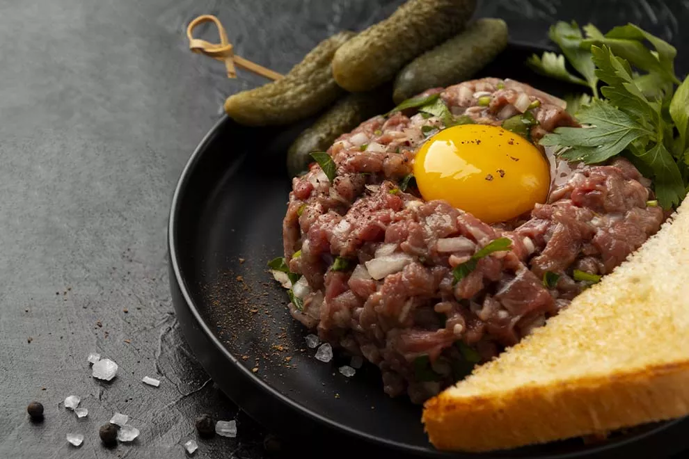 Imagem de um steak tartare com uma gema de ovo por cima, com salsinha e cebolinha picados, sob um prato redondo escuro, em cima de uma mesa 