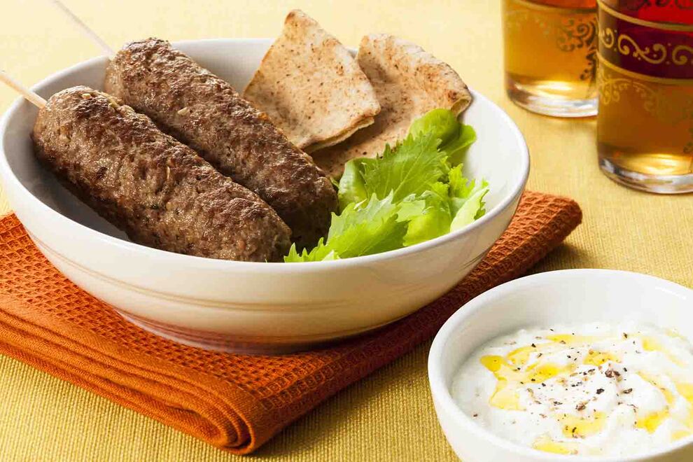 Imagem de duas unidades de espetinho da Kafta, em uma tigela redonda de cerâmica, com fatias de pão sírio e folhas de salada, sob uma mesa com toalha amarela, com uma tigela pequena com húmus ao lado 