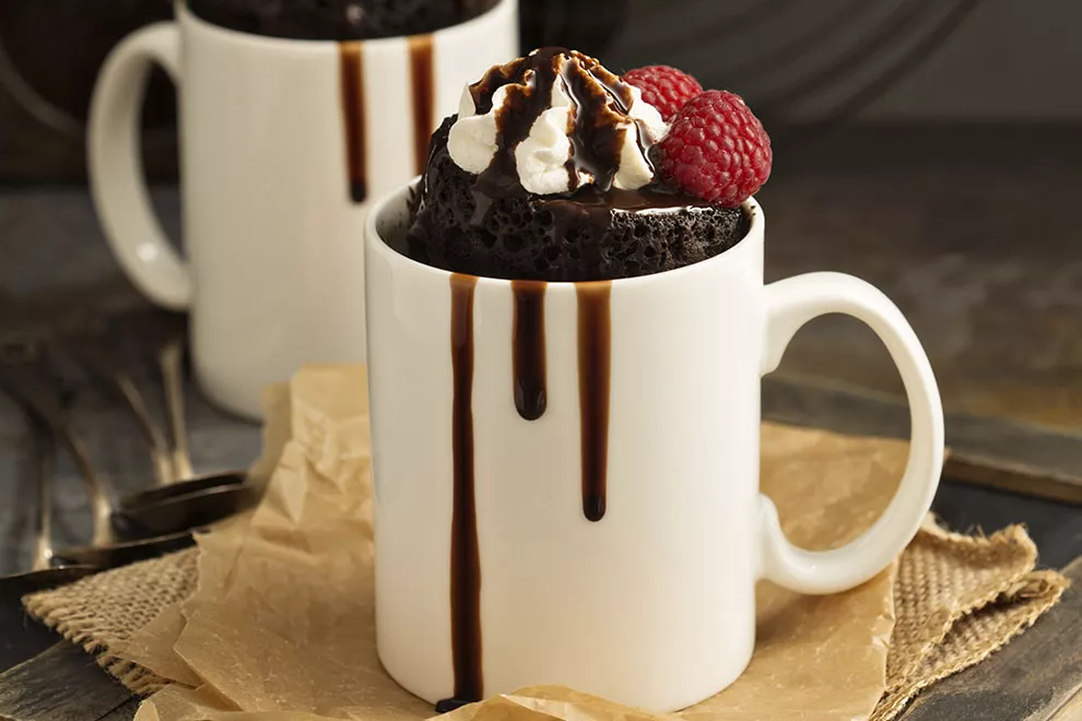 Imagem de um bolo de chocolate de caneca, com chantily , duas framboesas e calda de chocolate, em uma caneca branca, sob a mesa 