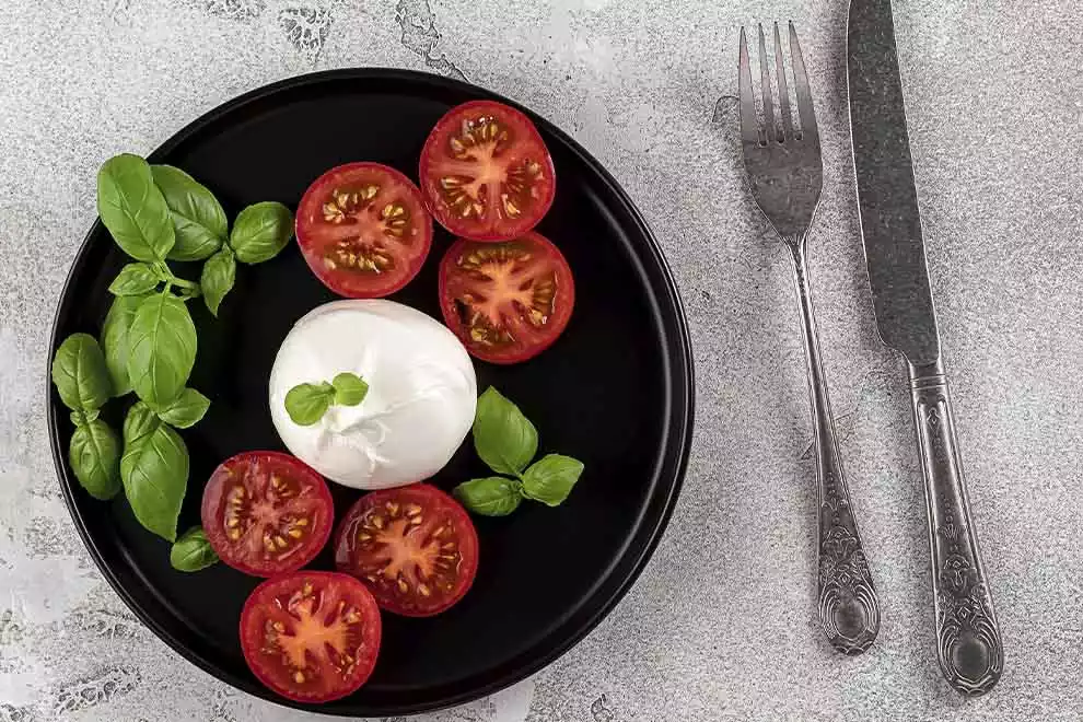 Imagem de um prato escuro com burrata, fatias de tomate e folhas de manjericão, sob uma mesa cinza com um par de talheres ao lado