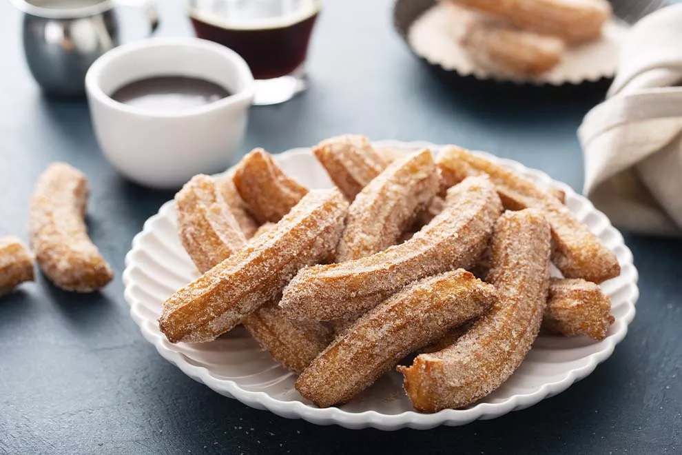 Imagem de diversos churros caseiros em cima de um prato, com uma xícara de café ao fundo, sob uma mesa escura.