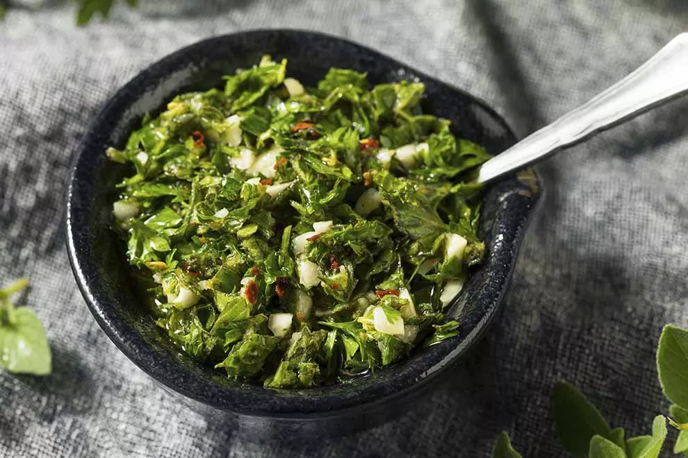 Chimichurri em uma pequena tigela de cerâmica, com uma colher nele, sob uma mesa de mármore.