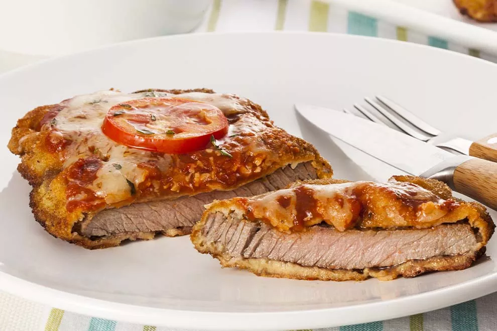 Bife à Parmegiana de contrafilé, cortado ao meio, em cima de um prato redondo branco, com um par de talheres ao lado,  sob uma mesa de madeira. 