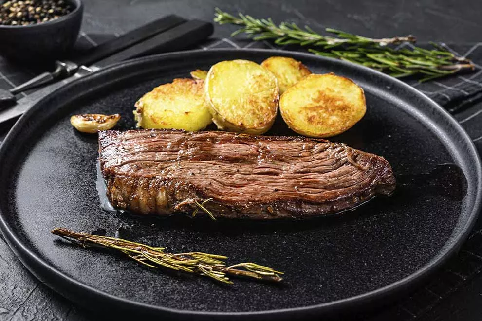 Imagem de uma fatia de fraldinha feita no forno, com batatas assadas e um ramo de alecrim ao lado, sob um prato escuro, em cima de uma mesa escura 