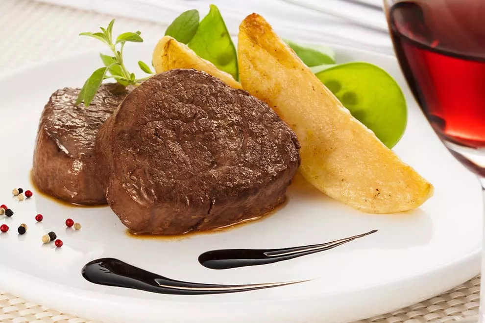 Imagem de um prato branco com duas unidades de filé mignon assados, com duas unidades de batata frita e folhas de salada ao lado, sob uma mesa, com uma taça de vinho no canto da imagem. 