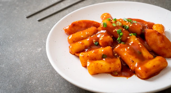 Tteokbokki servido em um prato branco, redondo e raso. Ao fundo, um par de hashis.