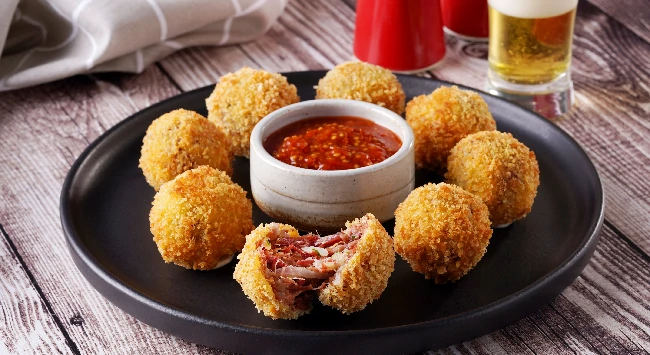 Bolinhos de mandioca recheados com carne seca em um prato preto. Ao fundo, um copo com cerveja.