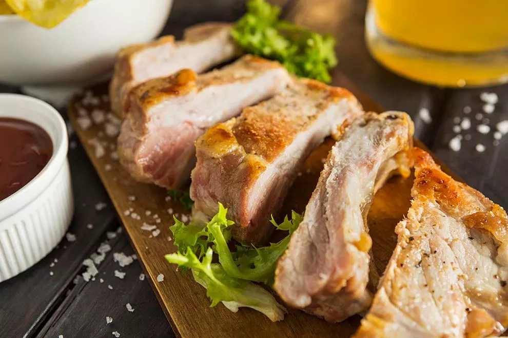 carne de porco bem assada em cima de uma tábua de madeira com um pote de molho ao lado e em cima de uma mesa de madeira escura